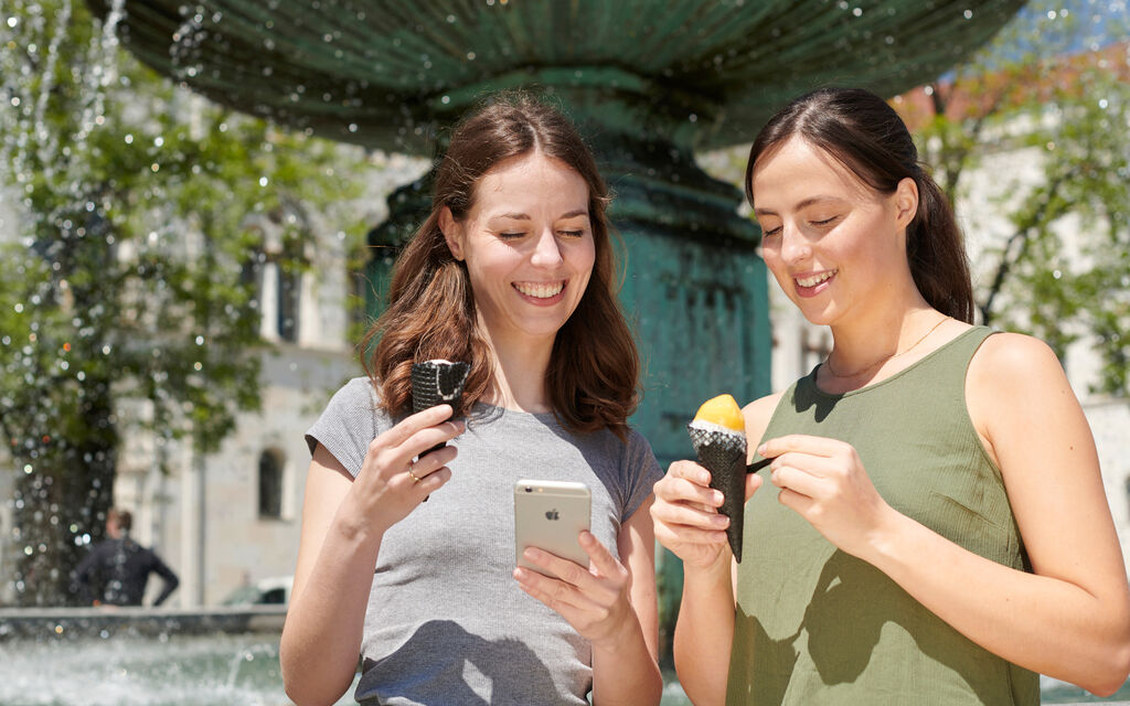 Testing the M-Login Two young women standing in front of a fountain, eating ice cream and looking at a smartphone.
