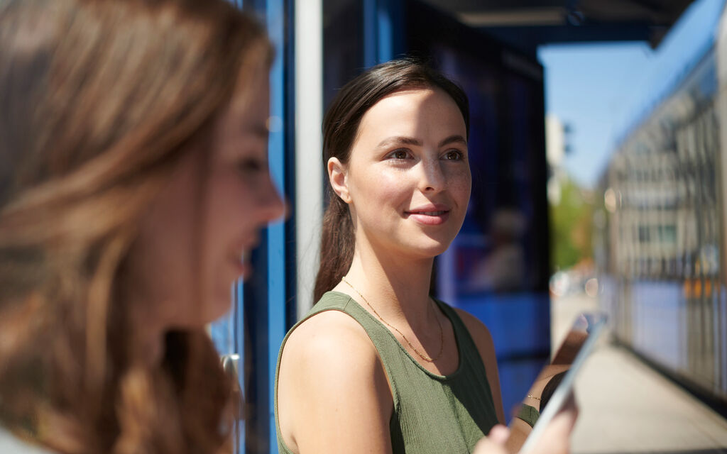 Testing the M-Login Young woman sitting at a tram station, in the foreground a woman looking at her smartphone