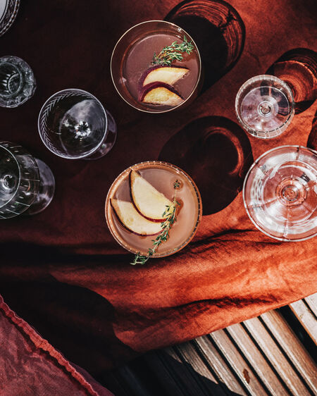 KPM glasses, two of them filled with a peach summer drink, on rust-coloured picnic blanket on jetty
