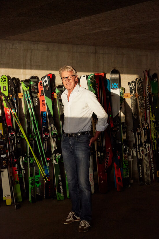 Thrill is his job: How Bernhard Russi develops ski slopes for top-class sport Bernhard Russi stands in front of a wall with several skis and ski poles.