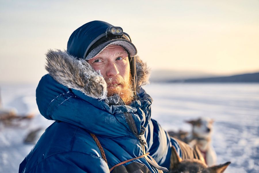 Dog sledding in Lapland: Visiting a husky farm Portrait photo of dog sledge driver Jonas in blue winter coat with fur collar at sunset