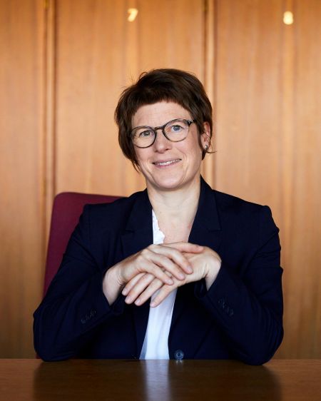 Veronika Grimm in a suit sitting in front of a wooden wall