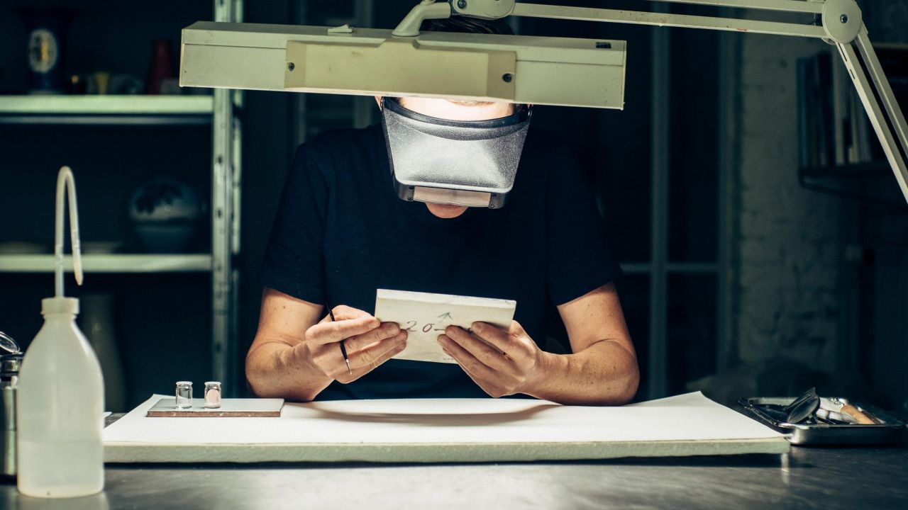 A restorer with protective glasses sits at a workbench, examining a white ceramic object under a desk lamp A restorer with protective glasses sits at a workbench, examining a white ceramic object under a desk lamp