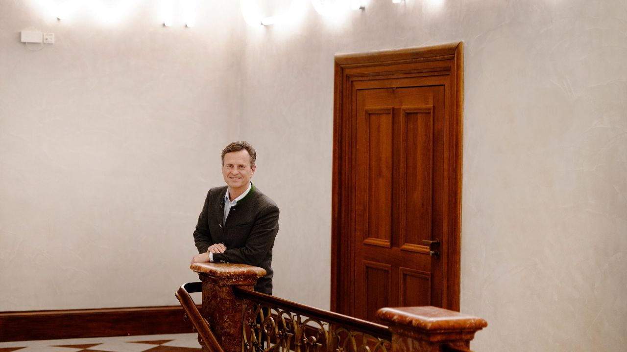 A smiling man stands at the staircase railing in a foyer, with neon letters spelling out "Wirtschaft" glowing in the background. A smiling man stands at the staircase railing in a foyer, with neon letters spelling out "Wirtschaft" glowing in the background.