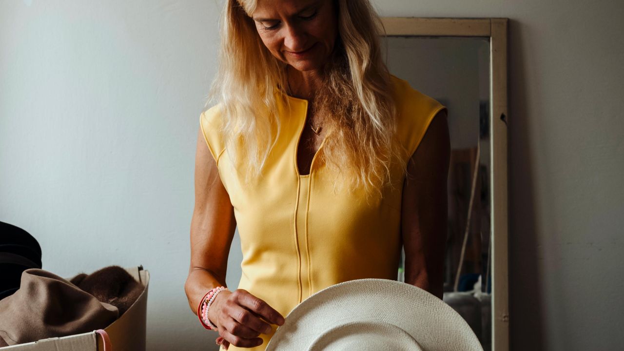A blonde woman in a yellow dress holds a light-colored hat in front of her body with both hands. A blonde woman in a yellow dress holds a light-colored hat in front of her body with both hands.