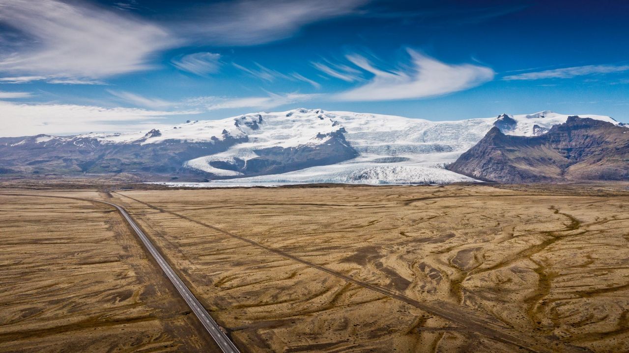 Luftaufnahme einer leeren Straße, die durch eine karge Ebene mit Gletscher und Bergen in Island führt. Luftaufnahme einer leeren Straße, die durch eine karge Ebene mit Gletscher und Bergen in Island führt.