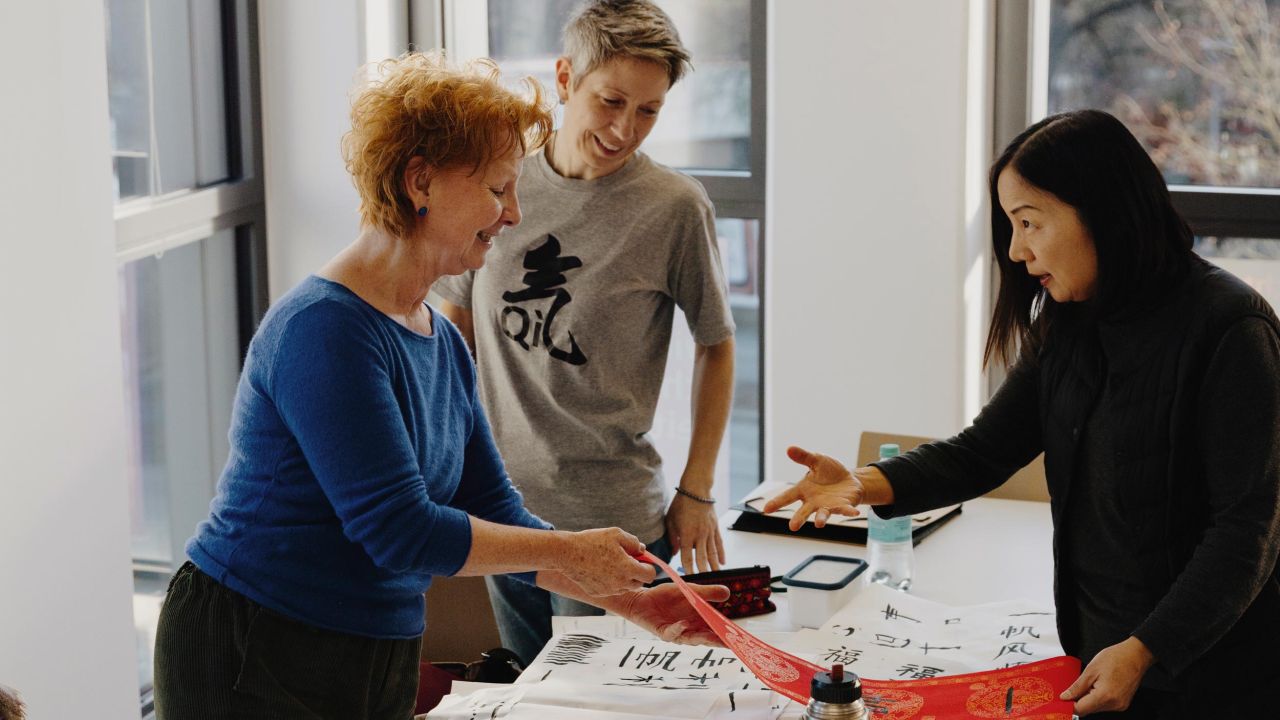Three women are standing at a table in a bright room, smiling and discussing calligraphy work. Three women are standing at a table in a bright room, smiling and discussing calligraphy work.