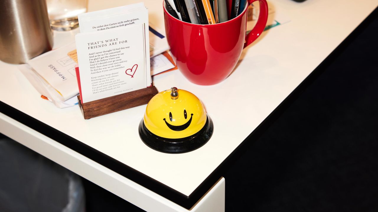 A yellow and black bell with a smiley face is placed on a white desk next to a business card holder and a red pen holder A yellow and black bell with a smiley face is placed on a white desk next to a business card holder and a red pen holder